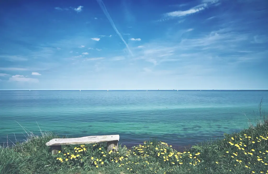 Ruhiges Meer unter hellem Himmel, vorn eine Bank in den Dünen