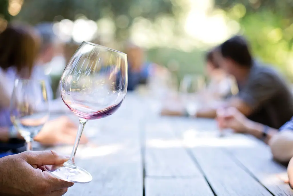 Gäste an einem langen Holztisch im Freien, im Vordergrund ein Glas Rotwein