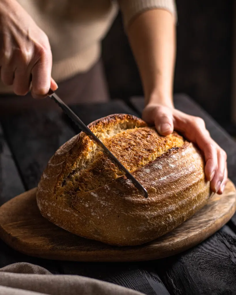 Hände schneiden ein dunkles Sauerteigbrot auf einem Holzbrett an